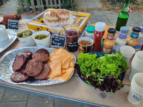 Een schaal met gegrild vlees, brood en salades staat klaar op
                              een tafel bij het buurtfeest.