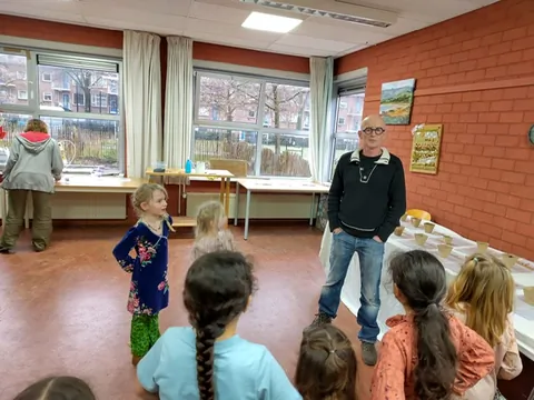 Kinderen zitten in een buurtruimte te luisteren naar een
                              begeleider die uitleg geeft.