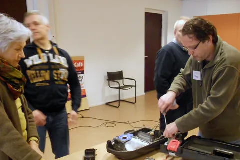 Twee bezoekers van het repaircafé buigen zich over een apparaat
                              dat op tafel wordt gerepareerd.