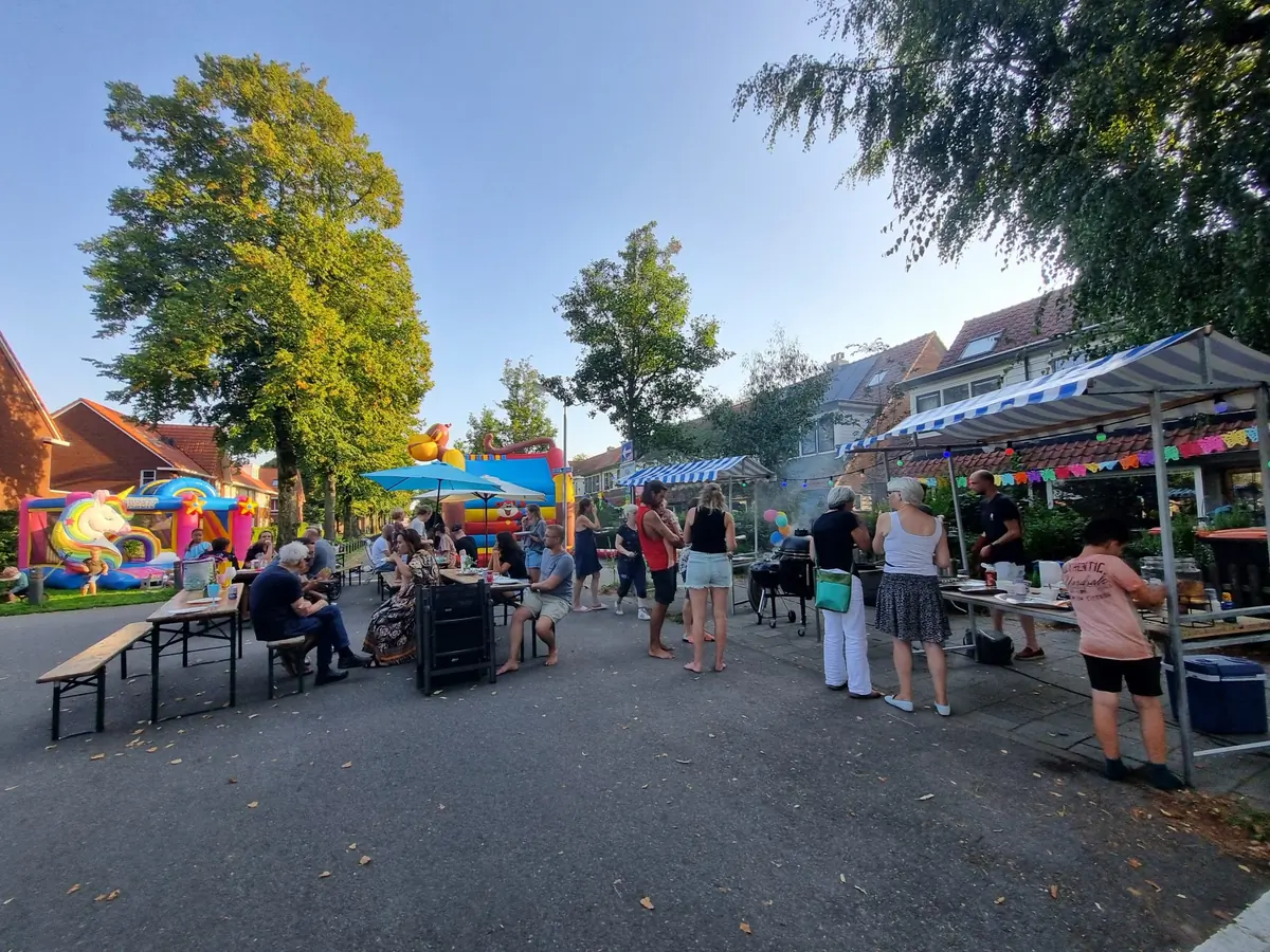 Buurtbewoners staan in de avondzon bij kraampjes en partytenten op een
                      afgesloten straat tijdens een buurtfeest.
