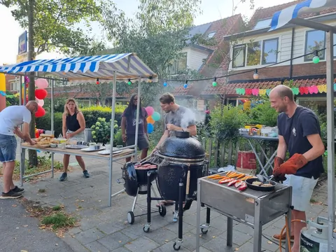 Twee buurtbewoners bakken vlees op barbecues midden op de straat,
                              met slingers tussen de huizen.