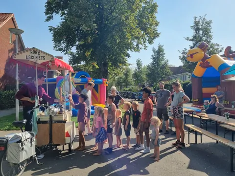 Kinderen staan in de rij bij een ijskar op een zonnige straat,
                              met picknicktafels en een luchtkussen op de achtergrond.