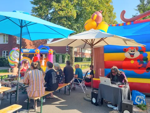 Buurtbewoners zitten onder blauwe parasols aan picknicktafels
                              naast een groot luchtkussen.